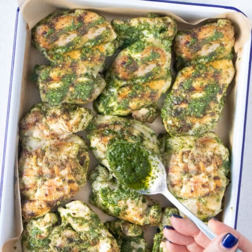 A white hand is spooning bright green cilantro chimichurri over pan seared chicken thighs.