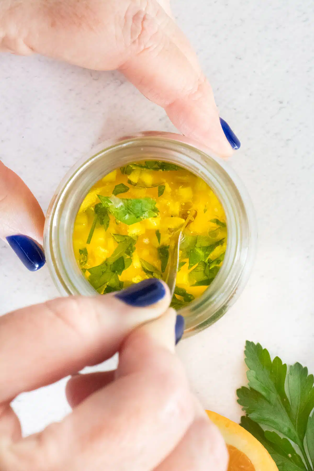 A white hand with blue nail polish is stirring the vinaigrette in a small jar.