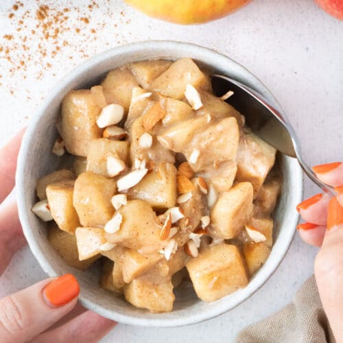 Two white hands are holding a small bowl filled with stewed apples topped with chopped almonds.