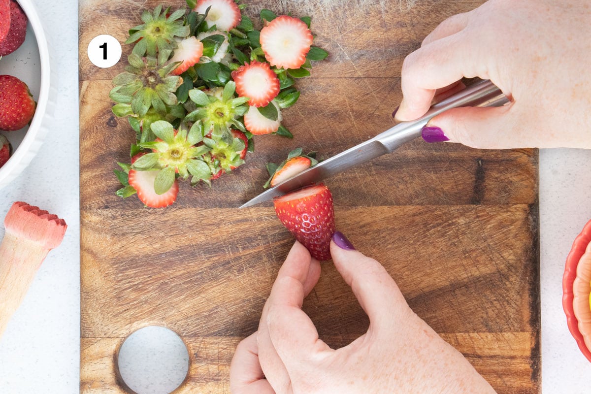 A white hand is holding a bright red strawberry on a wooden board while the other hand chops off the top with a knife.