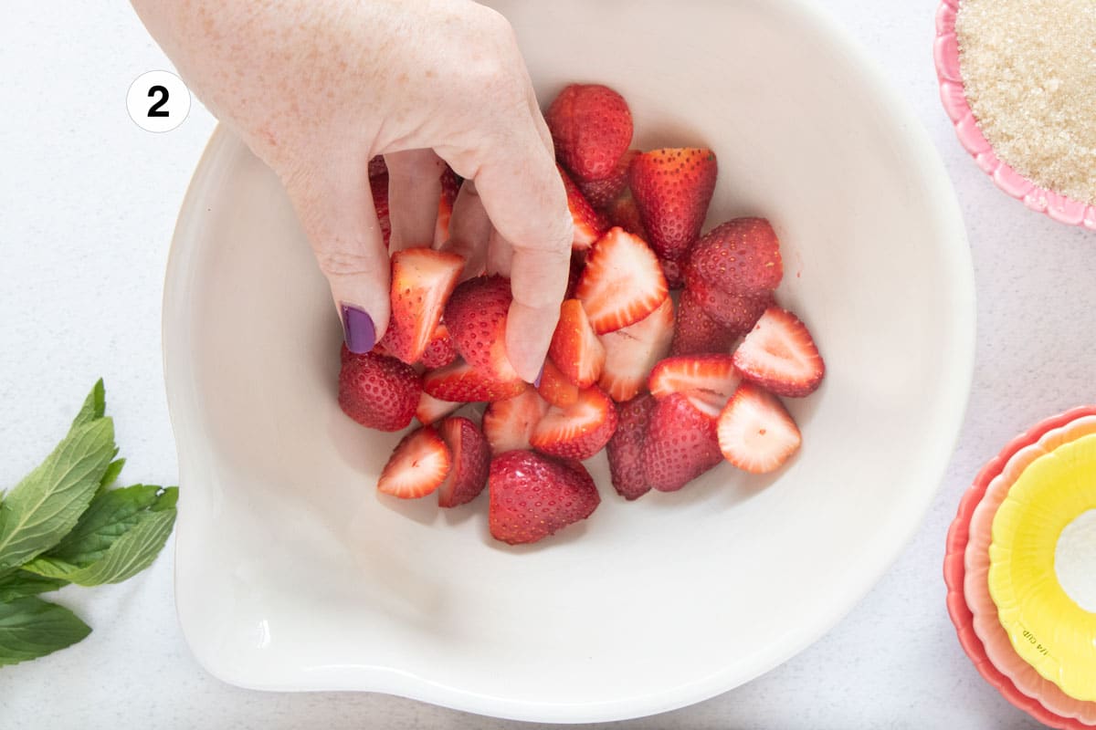 A white hand is dropping bright red strawberry halves into a large cream-coloured bowl.