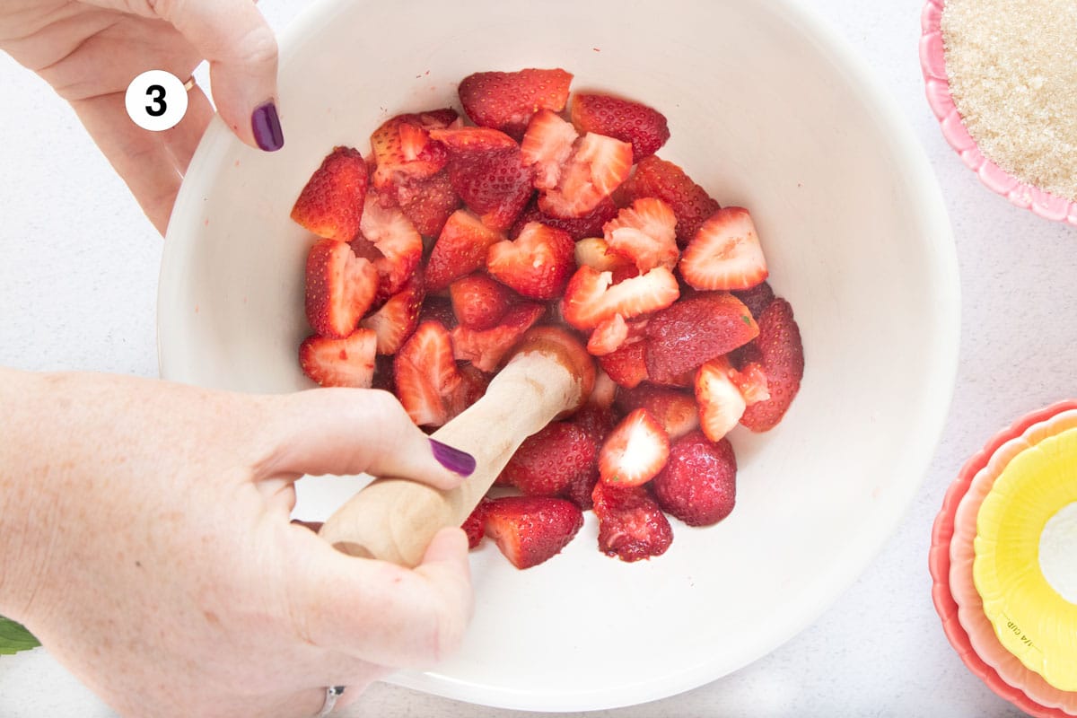 A white hand is using a wooden muddler to crush bright red strawberries in a large cream-coloured bowl.