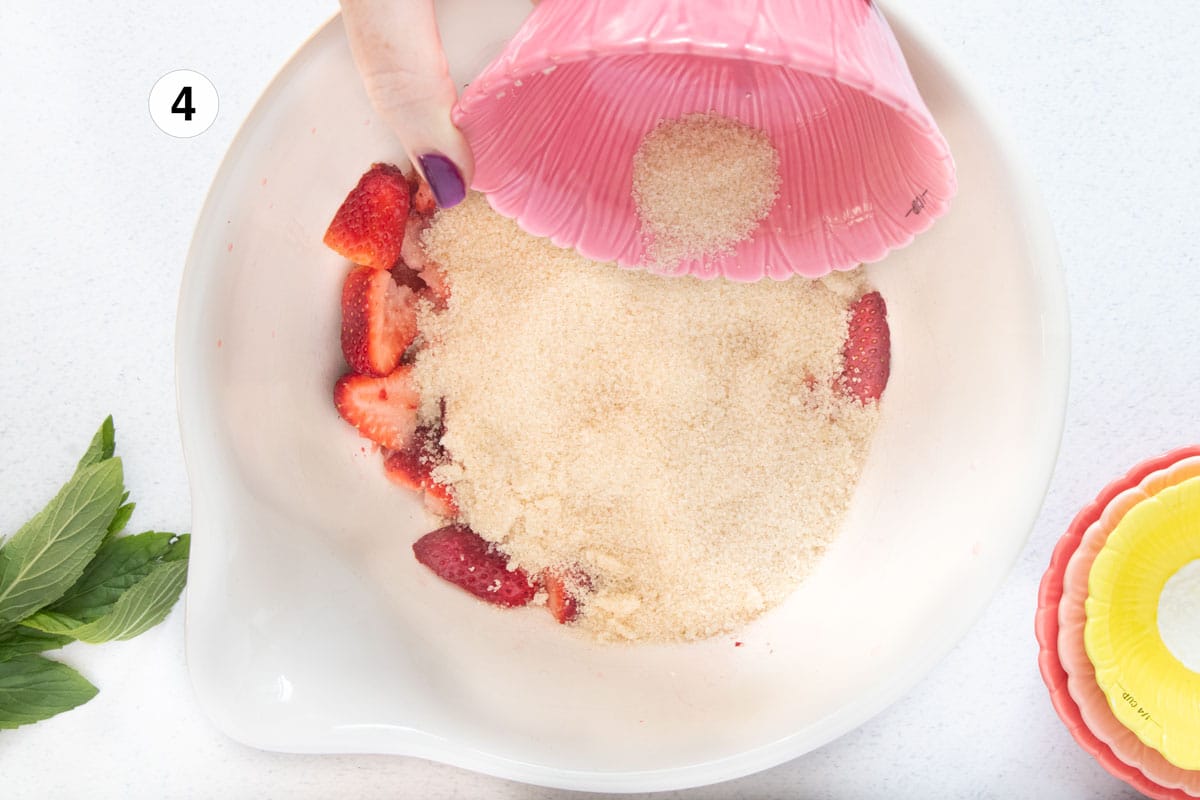 Raw sugar is being poured over strawberries in a large bowl to make the base for a strawberry shrub.