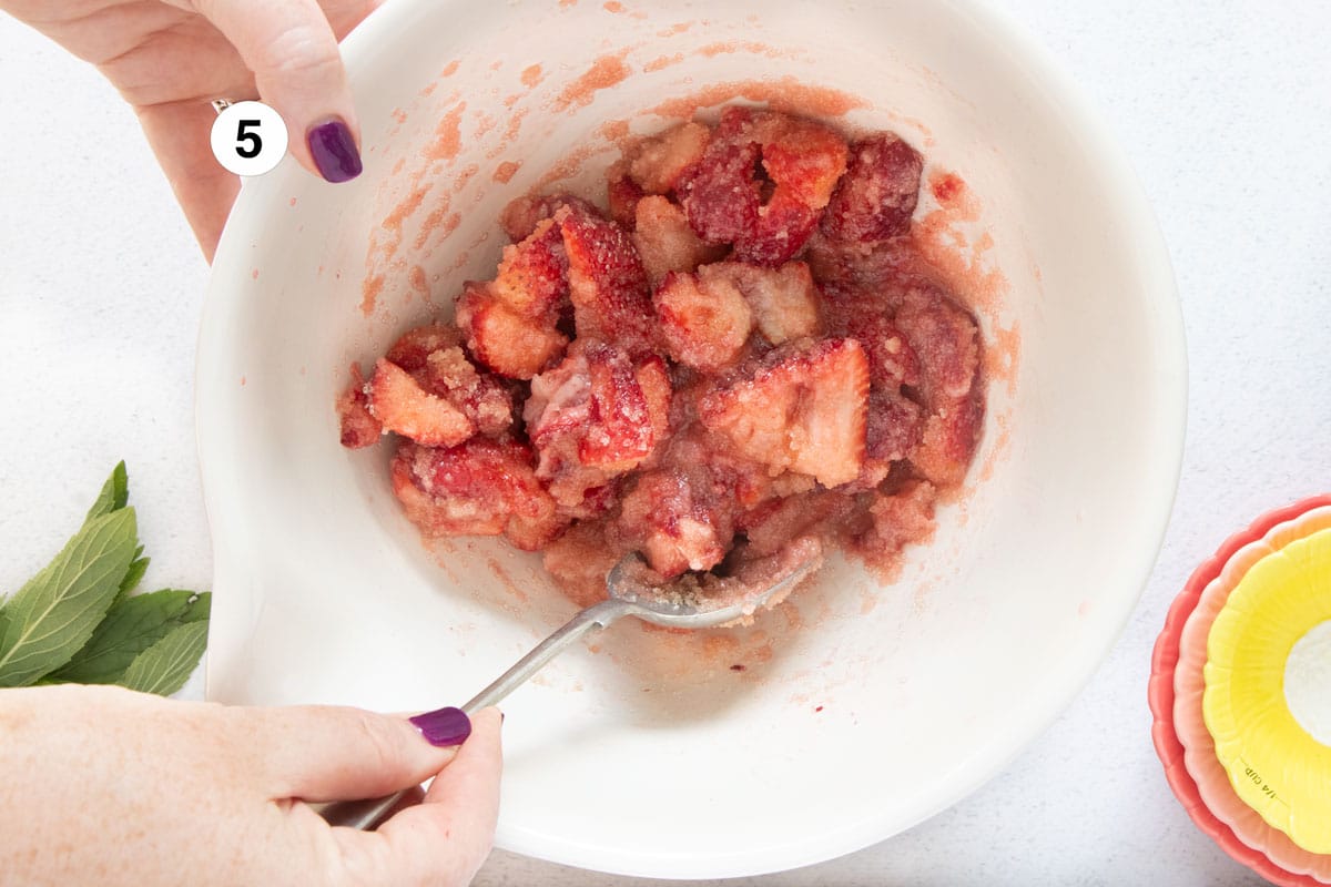A white hand is stirring sugar and strawberries together in a bowl with a silver spoon.
