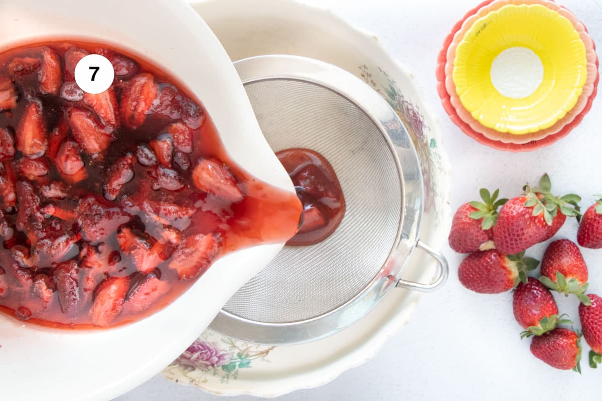 A large bowl of strawberry shrub is being poured through a fine mesh sieve into another bowl to catch the syrup.