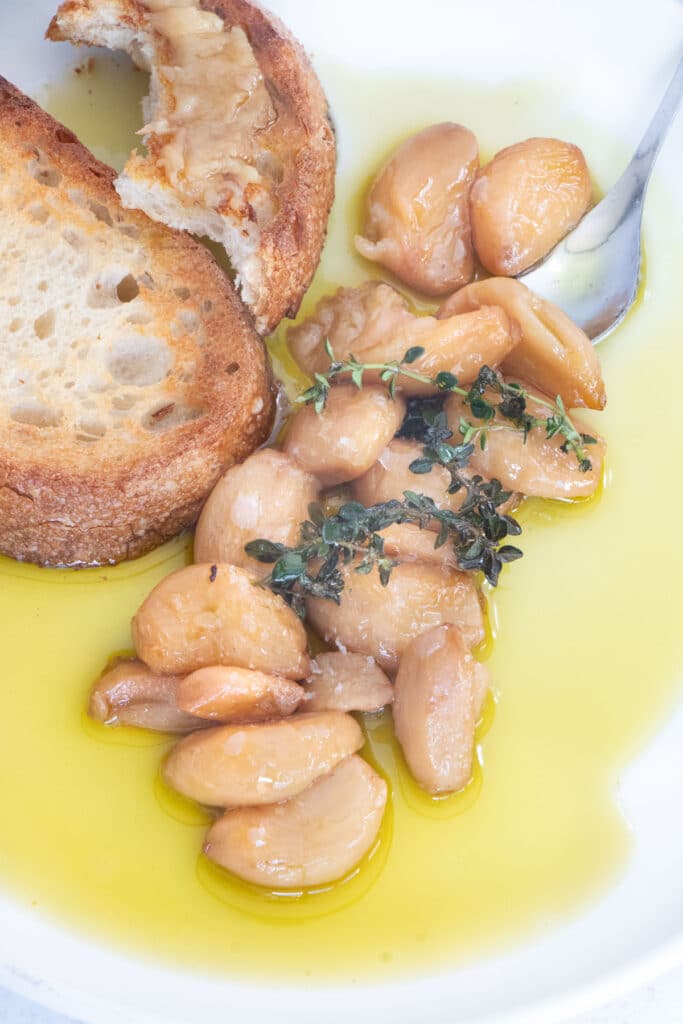 Cloves of confit garlic are piled on a plate drizzled with oil. A piece of toast on the side has been spread with the garlic.