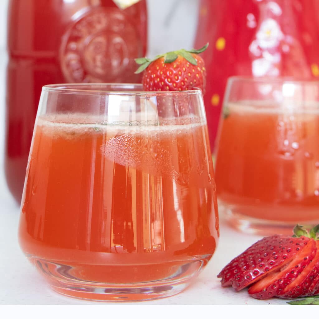 Two glasses of pink strawberry shrub are sitting side by side on a table. A bottle of shrub is behind them.