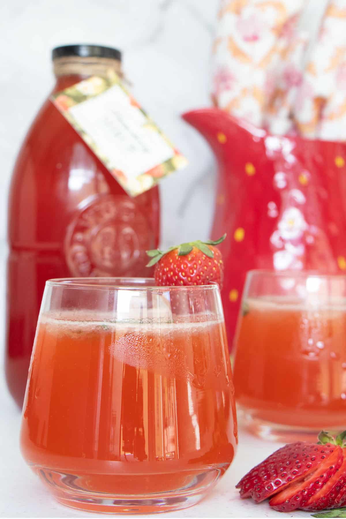 Two glasses filled with strawberry shrub and mineral water are sat on a table in front of a bottle of shrub.