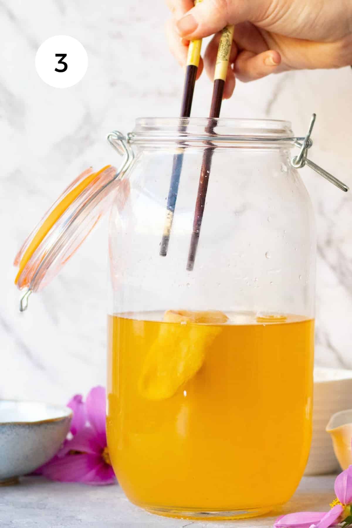 A white hand is dipping two chopsticks into a large jar of jun tea to take the scoby out.