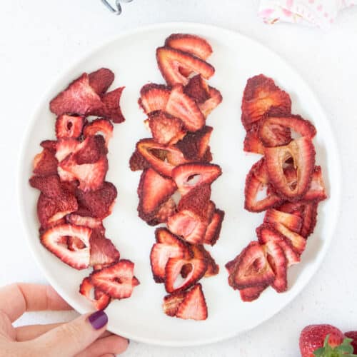 Three rows of bright red dehydrated strawberries are on a white plate being held by a white plate.