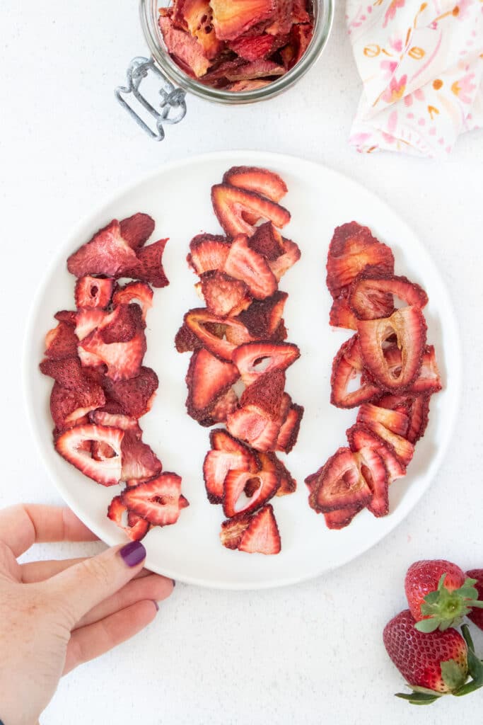 Three rows of bright red dehydrated strawberries are on a white plate being held by a white plate.