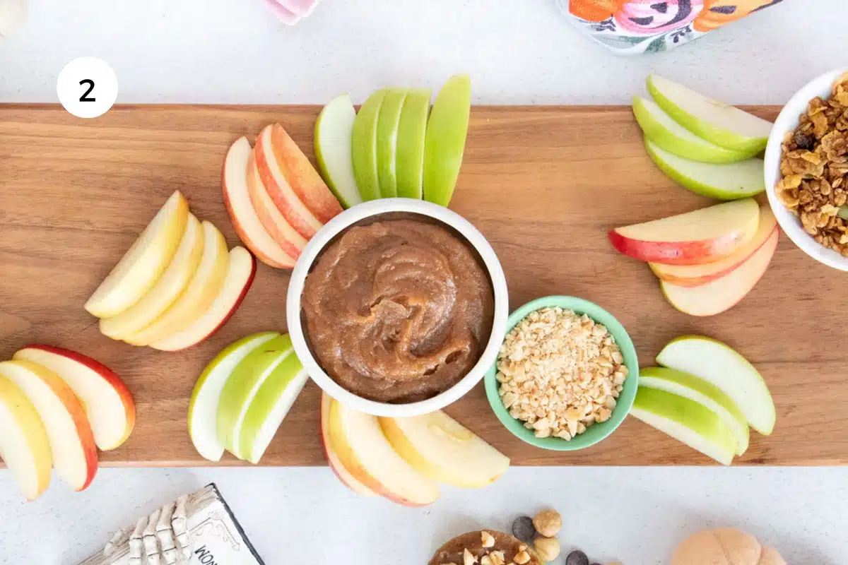 Small bowls full of toppings and sliced apples are starting to fill an apple dessert board.