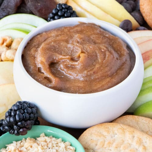 A white bowl filled with a thick date caramel sauce is sitting in the middle of a fruit, cookie, and nut platter.