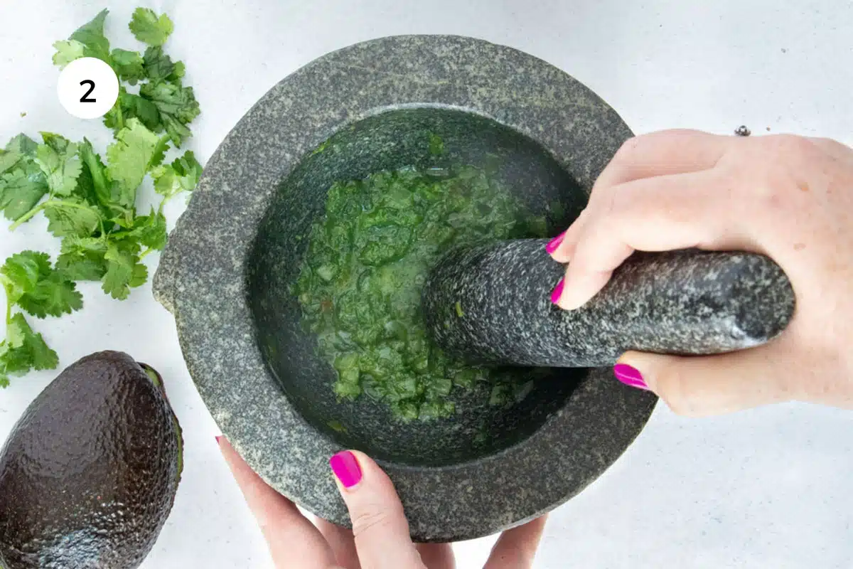 A white hand is holding a pestle and grinding ingredients in a mortar to make a green paste.