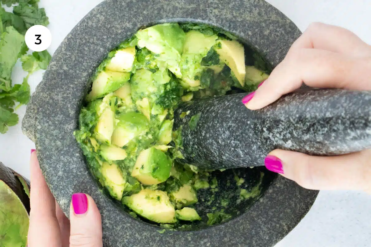 A white hand is mashing bright green avocado with a pestle in a large stone mortar.