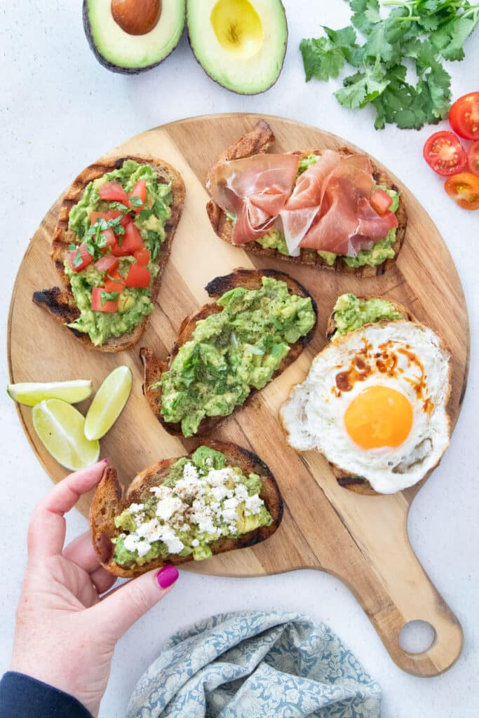 5 slices of guacamole toast with various toppings are laid out on a wooden serving board. A white hand is picking up a piece.