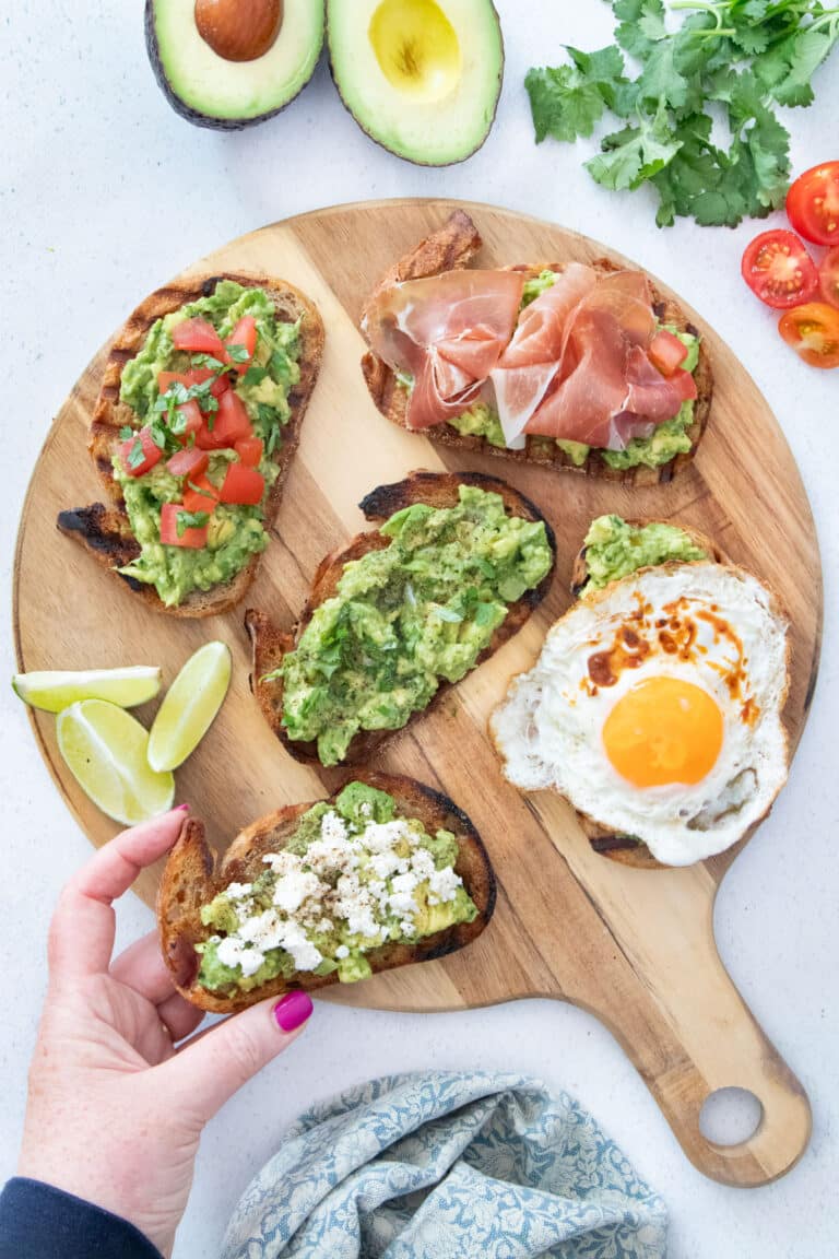 5 slices of guacamole toast with various toppings are laid out on a wooden serving board. A white hand is picking up a piece.