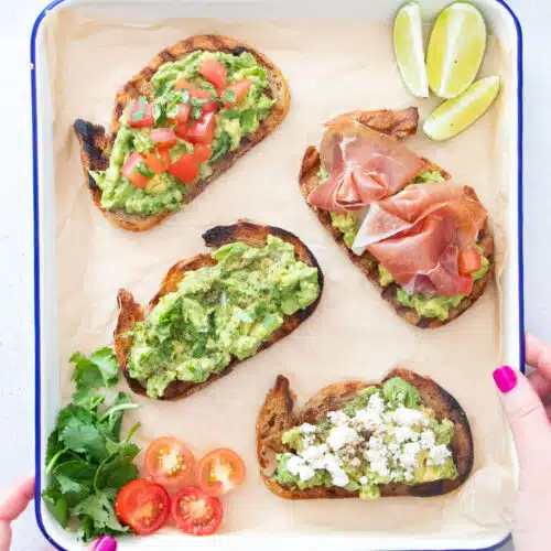 4 slices of guacamole toast with various toppings are laid out on a baking tray held by two white hands.