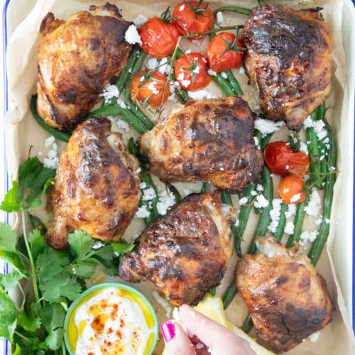 A white hand is sprinkling feta cheese over a cooked tray of Yogurt Marinated Chicken.