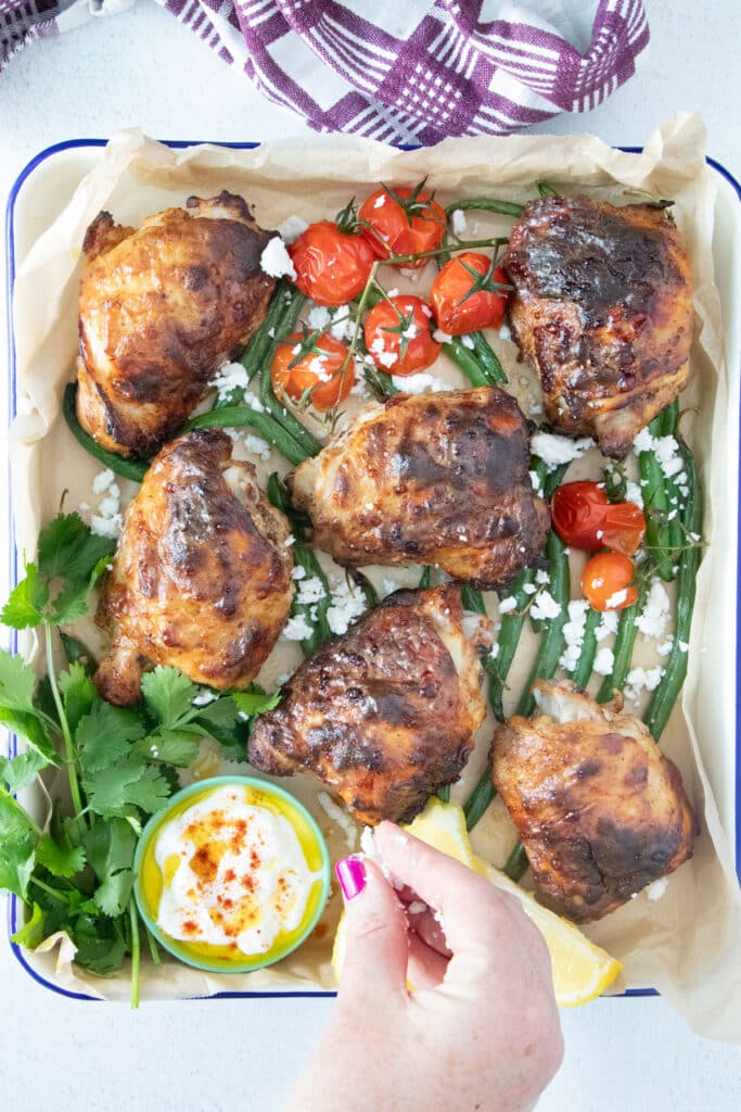 A white hand is sprinkling feta cheese over a cooked tray of Yogurt Marinated Chicken.