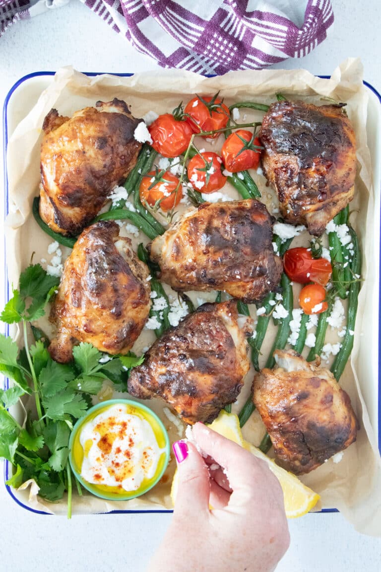 A white hand is sprinkling feta cheese over a cooked tray of Yogurt Marinated Chicken.