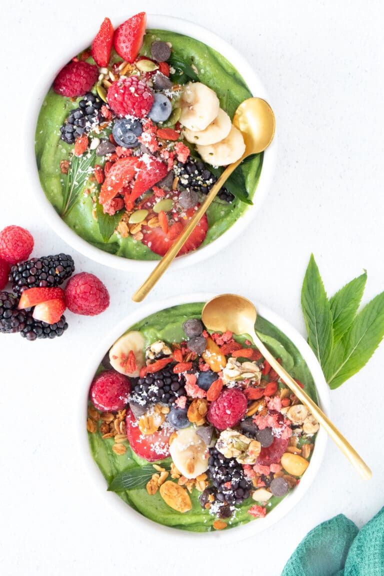 Two bright green smoothie bowls sit on a white table. They're topped with an assortment of berries and nuts.