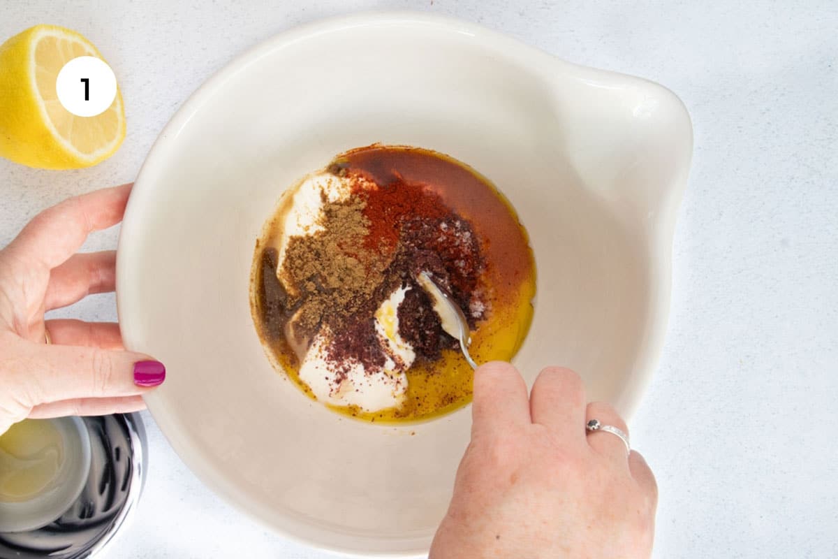 A white hand is using a silver spoon to mix the yogurt chicken marinade in a large bowl.