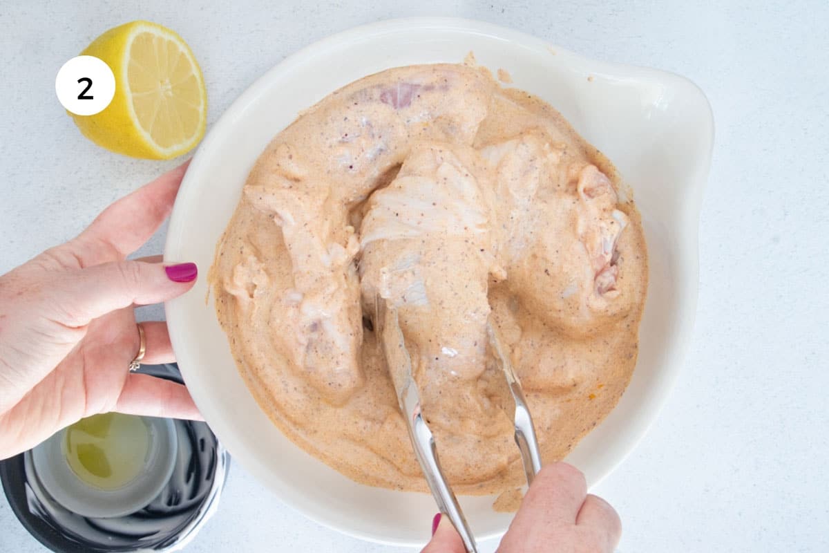 A white hand is using tongs to toss chicken thigh filler through a yogurt marinade in a large bowl.