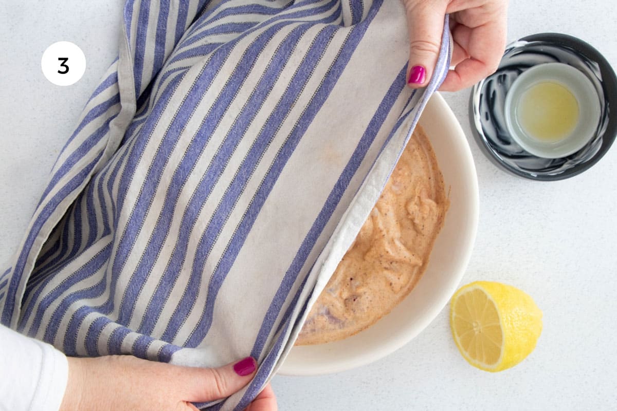 Two white hands are placing a blue and white striped tea towel over a bowl full of marinating chicken.