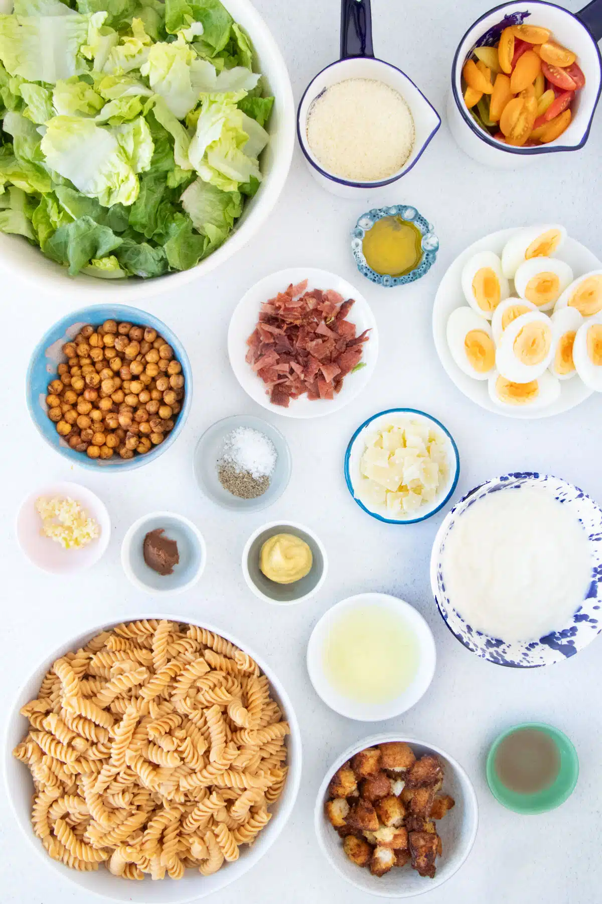 All the ingredients needed to make Caesar Pasta Salad are laid out across a table in bowls.