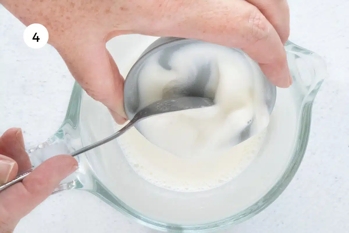 A white hand is tipping a small amount of yogurt into warmed milk in a jug to make a yogurt starter.