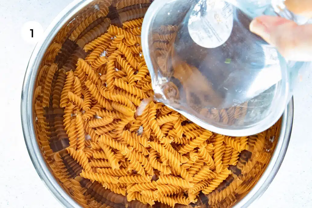 Pasta spirals are being added to an instant pot with water to cook for a Caesar Pasta Salad.