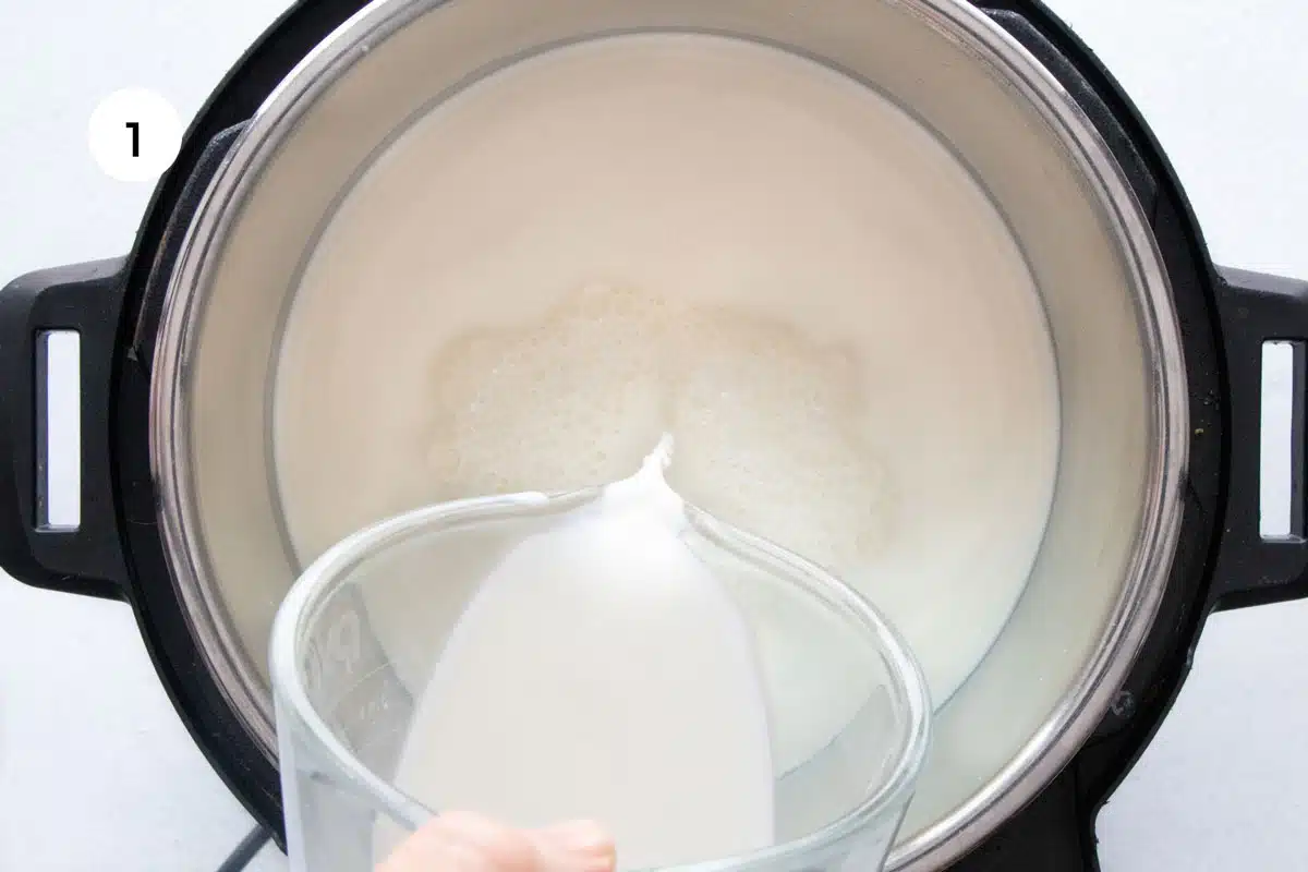 A white hand is pouring a jug of milk into the inner pot of an instant pot.