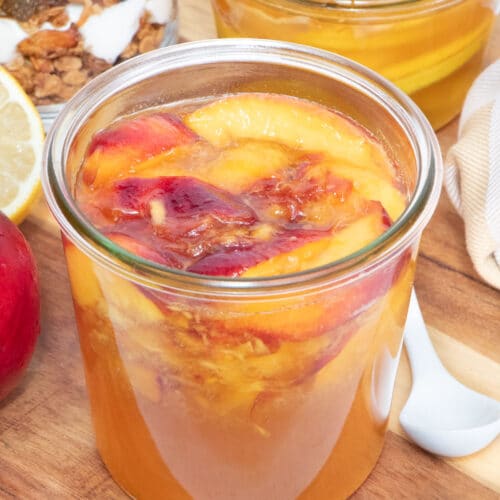 A jar of fermented fruit is sitting on a wooden serving board. Another of fermented lemon and ginger honey sits behind.