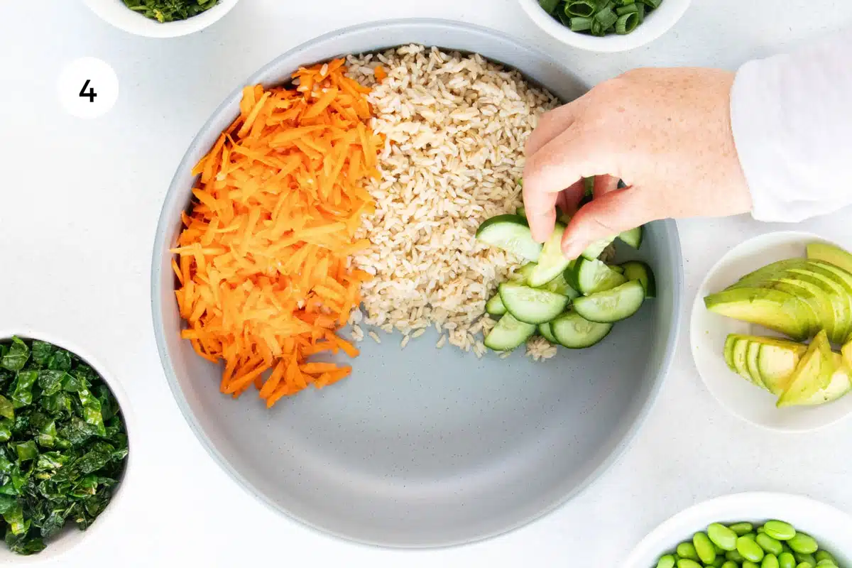 A white hand is piling ingredients into a large grey bowl to make an edamame salad.