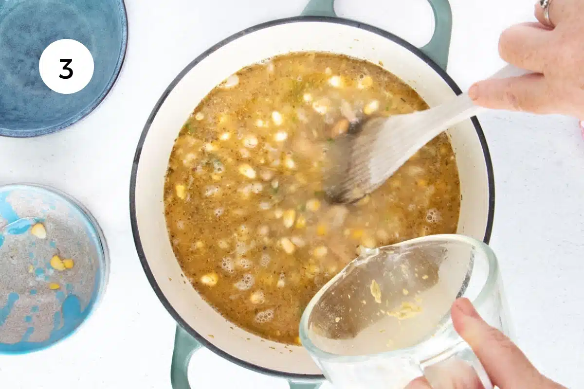 A white hand is pouring stock from a measuring cup into a large pot of vegetarian white chili.
