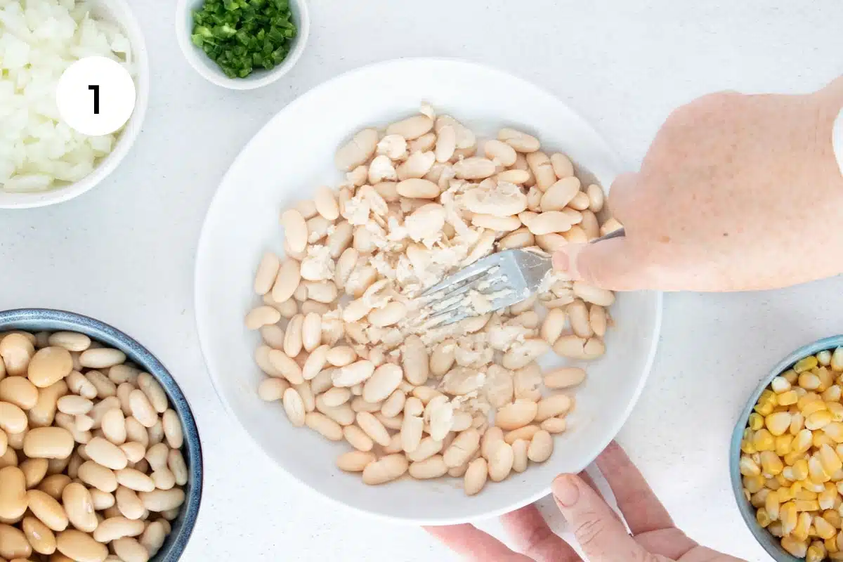 A white hand is using a silver fork to mash cannellini beans in a large white bowl on a tabletop.