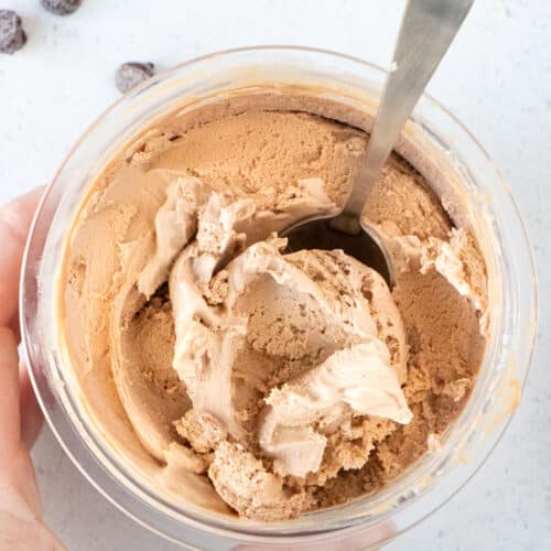 A white hand is holding a small Ninja Creami tub full of chocolate ice cream. A spoon is sticking out the top.
