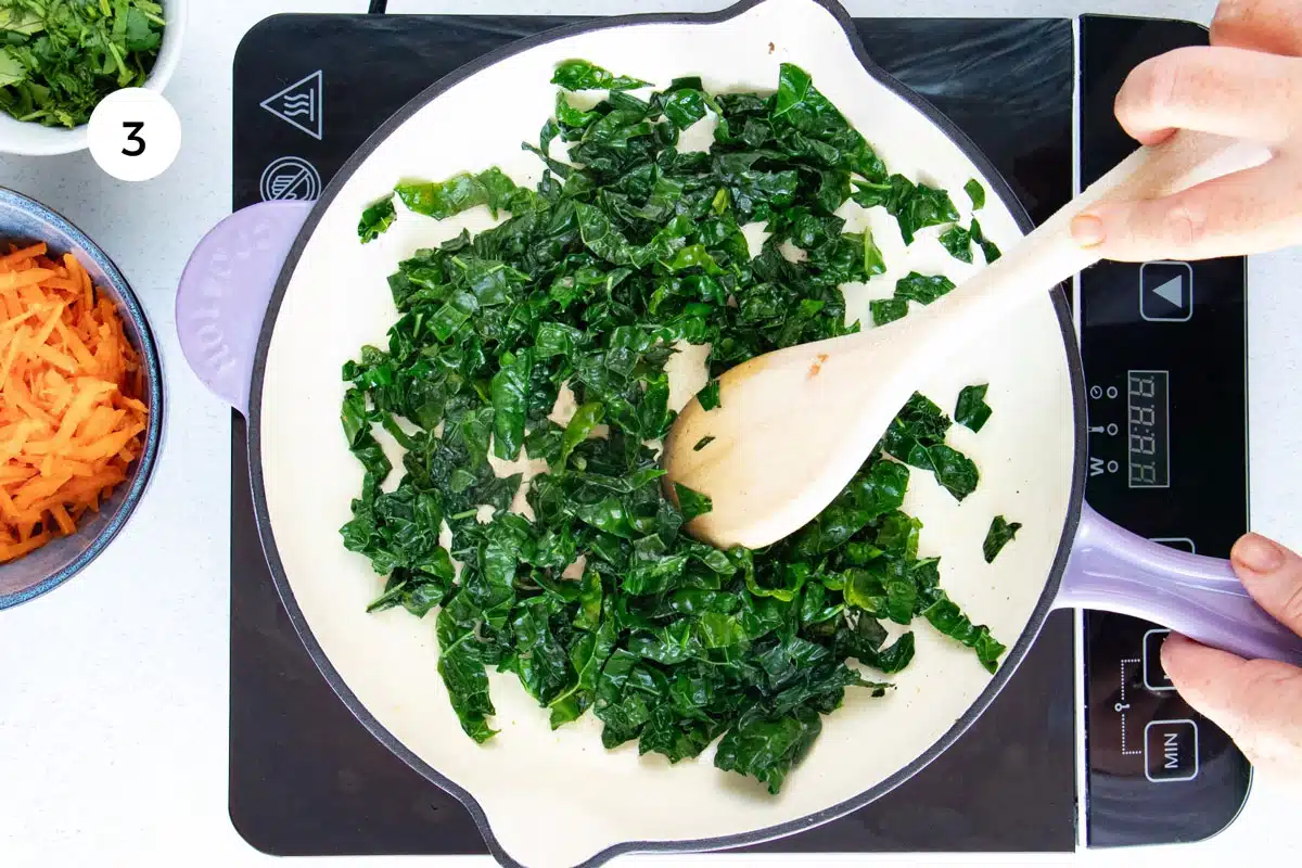Chopped kale is pan-frying in a large skillet over an induction cooktop. A white hand is stirring it with a wooden spoon.