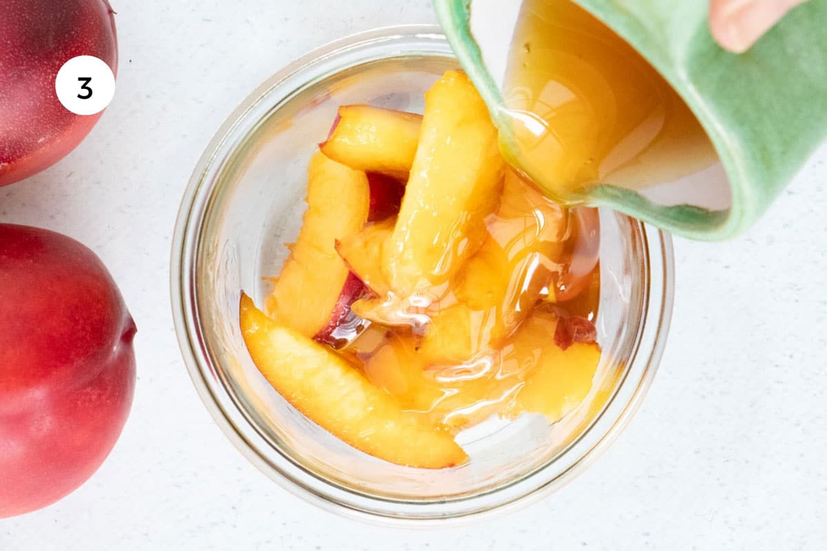 Honey is being poured from a green jug over slices of nectarine in a large glass jar.