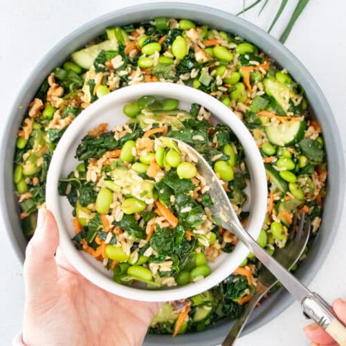 A white hand is holding a white bowl above a large edamame salad serving some into it.