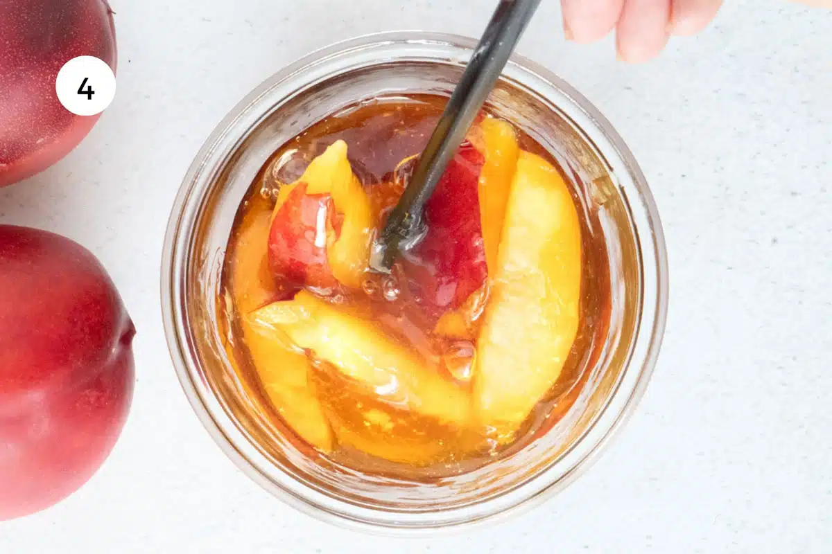 A white hand is using a chopstick to stir nectarine slices in honey in a large glass jar.