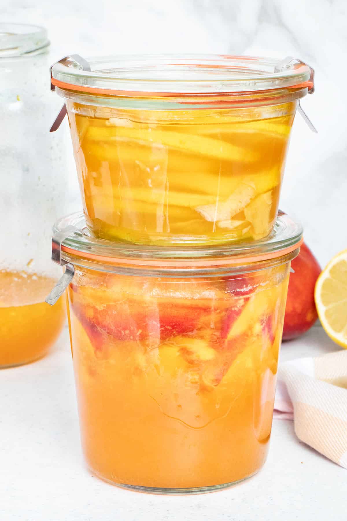 Two jars of honey fermented fruit are stacked on top of each other on a white bench-top.