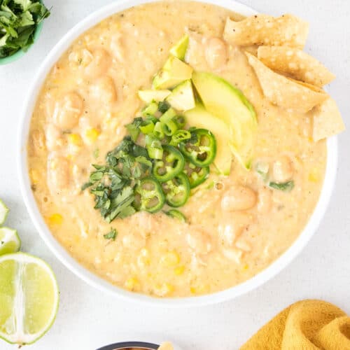 A large bowl of Vegan White Bean Chilli sits on a table top surrounded by a variety of toppings to be added.