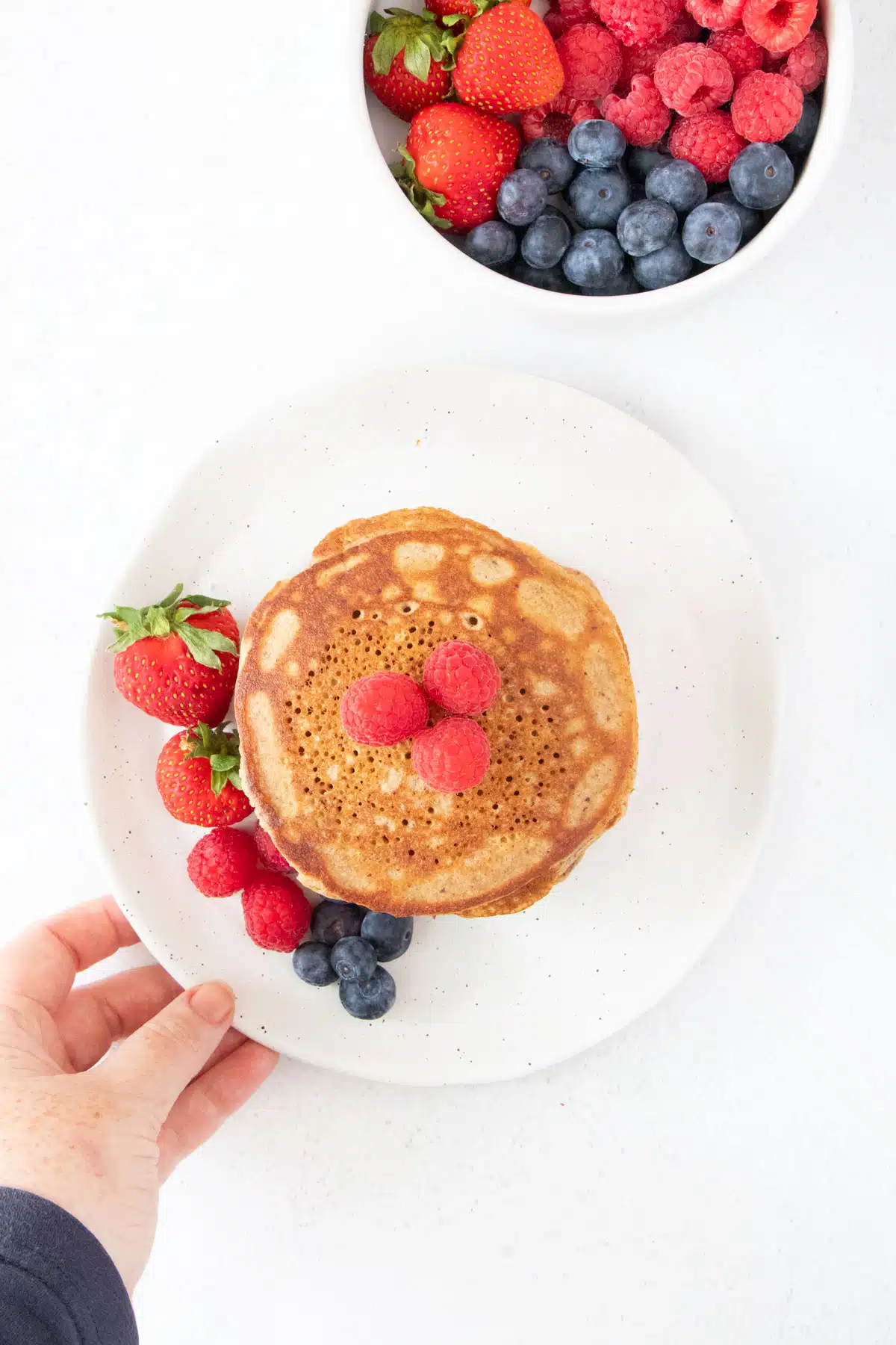 A white hand is holding a white plate filled with a stack of golden brown pancakes and mixed berries.