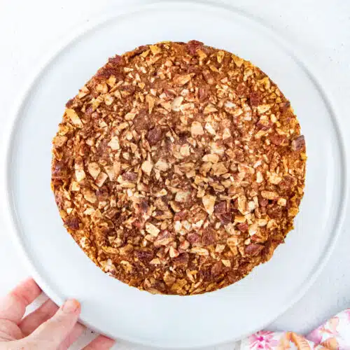 An almond and raspberry cake fresh out of the oven has been served on a white platter held by a white hand.