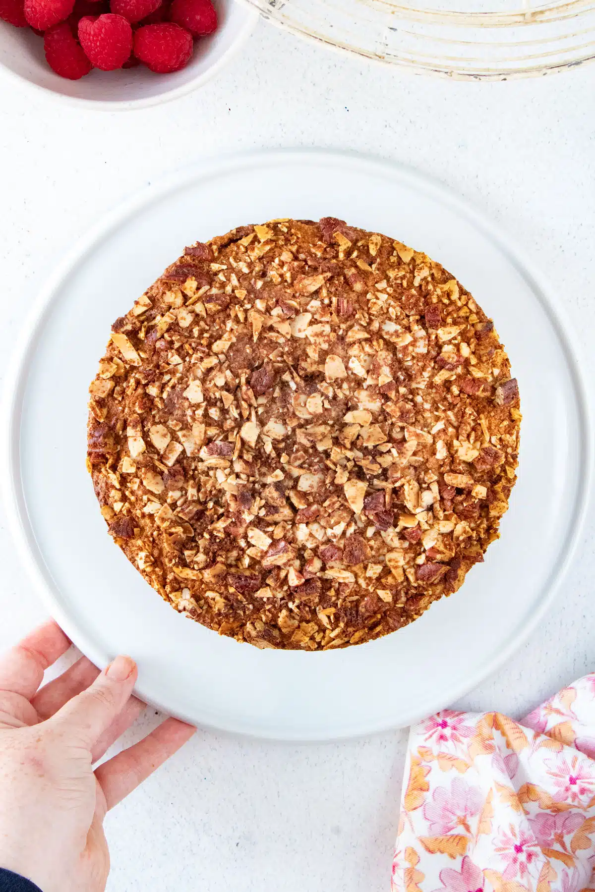 An almond and raspberry cake fresh out of the oven has been served on a white platter held by a white hand.