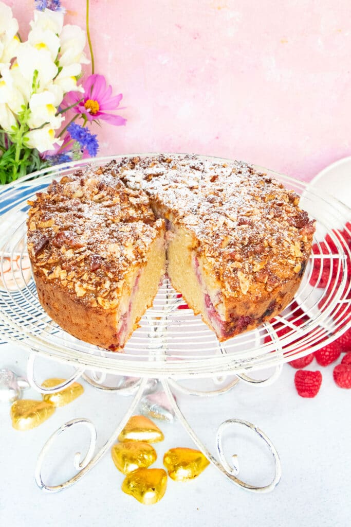 An almond and raspberry cake with a piece cut out is sitting on a white vintage cake stand.