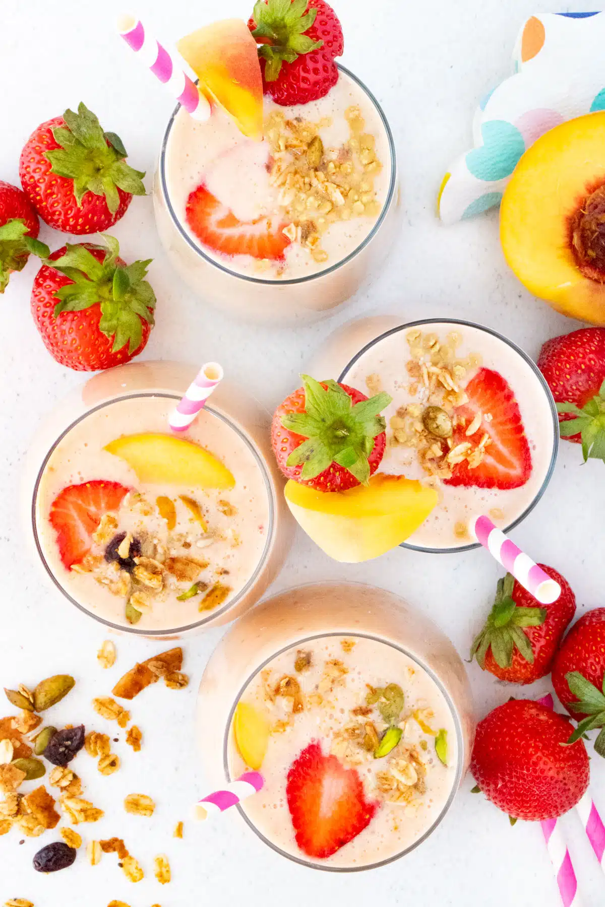 Four glasses are filled with strawberry and peach smoothie. Each is decorated with a strawberry plus a pink and white straw.