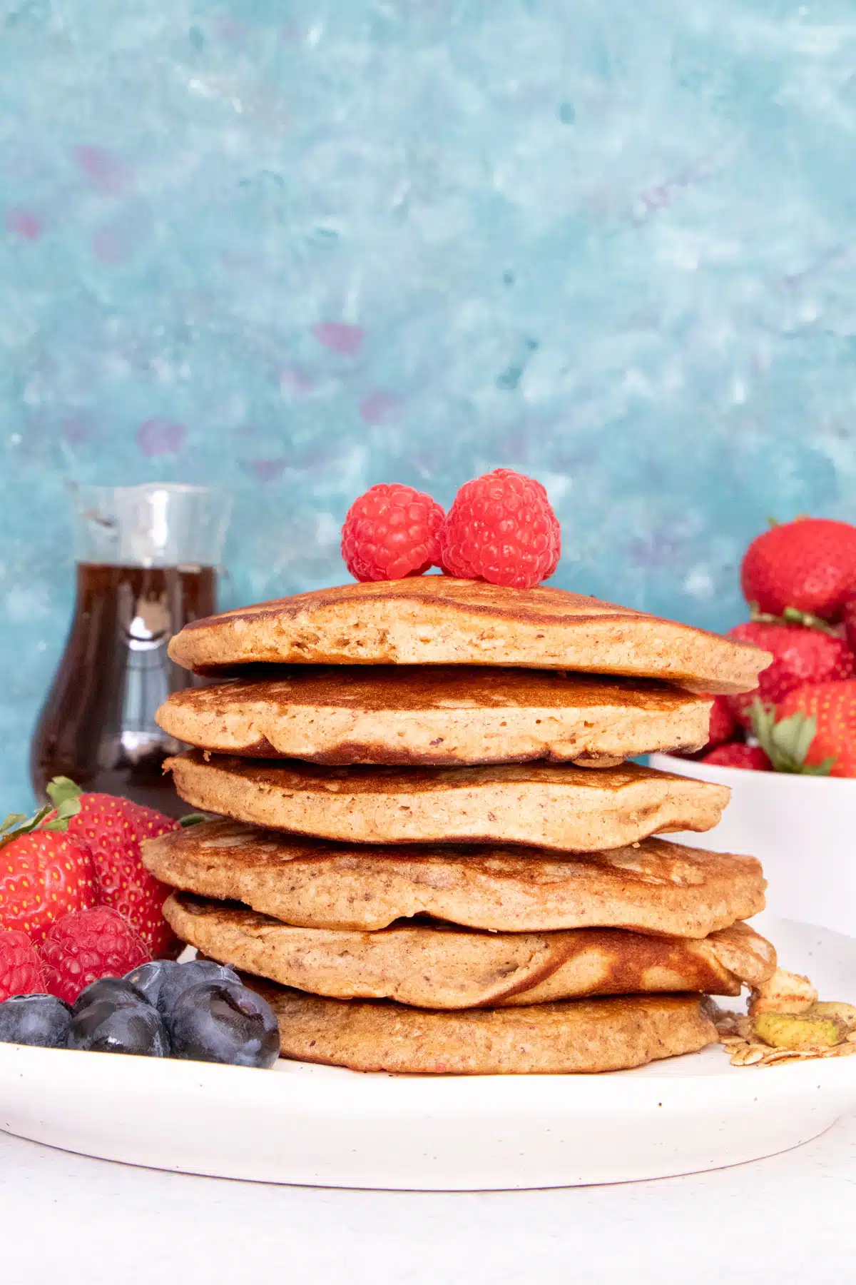 A large stack of golden brown pancakes on a plate with berries sits in front of a teal-coloured background.